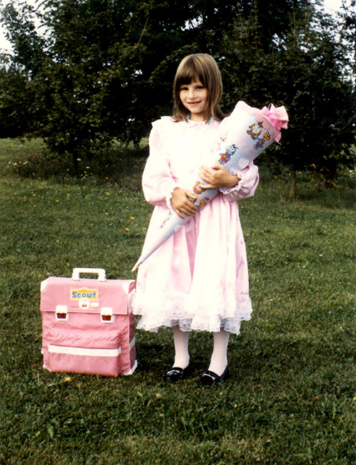 Mädchen im rosa Kleid bei seiner Einschulung mit Schultüte in der Hand und einem ebenfalls rosa Schulranzen neben sich.